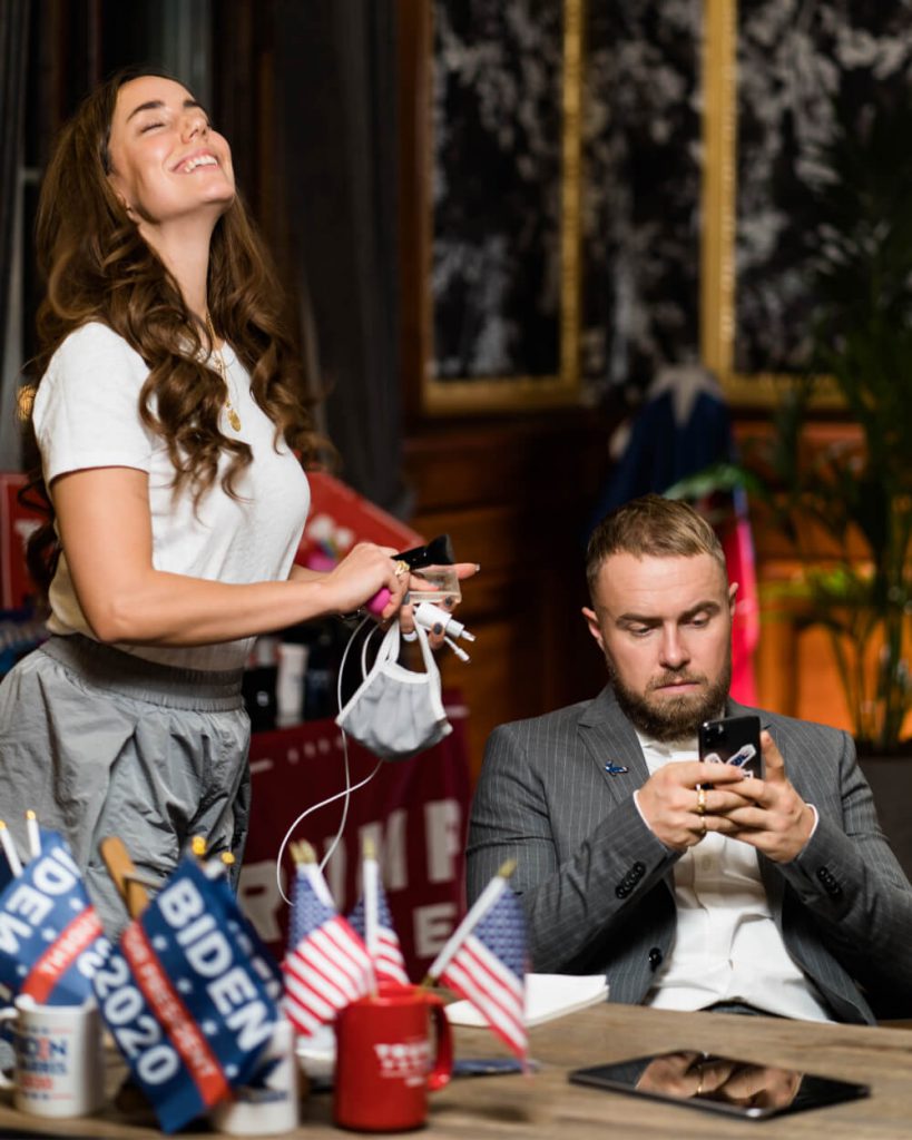 En mørkhåret kvinde står med et mundbind og en oplader ved siden af Peter Falktoft, som sidder ved et bord og kigger på sin telefon. I forgrunden ses flag med teksten "Biden 2020".