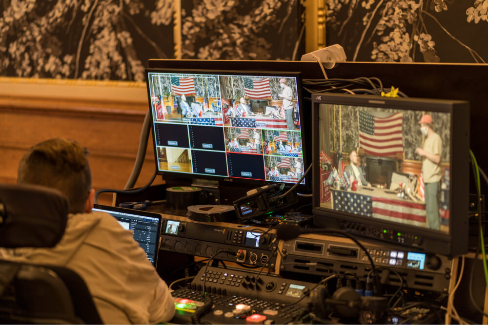 En mand sidder ved en mixerpult. På skærmen ses Esben Bjerre og en kameramand omgivet af kæmpestore amerikanske flag.
