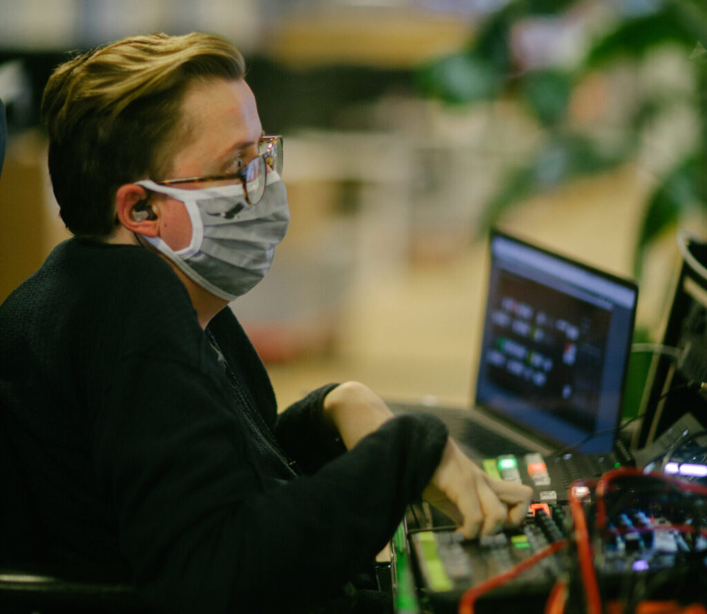 En mand iført mundbind sidder ved en mixerpult og producerer livestream.