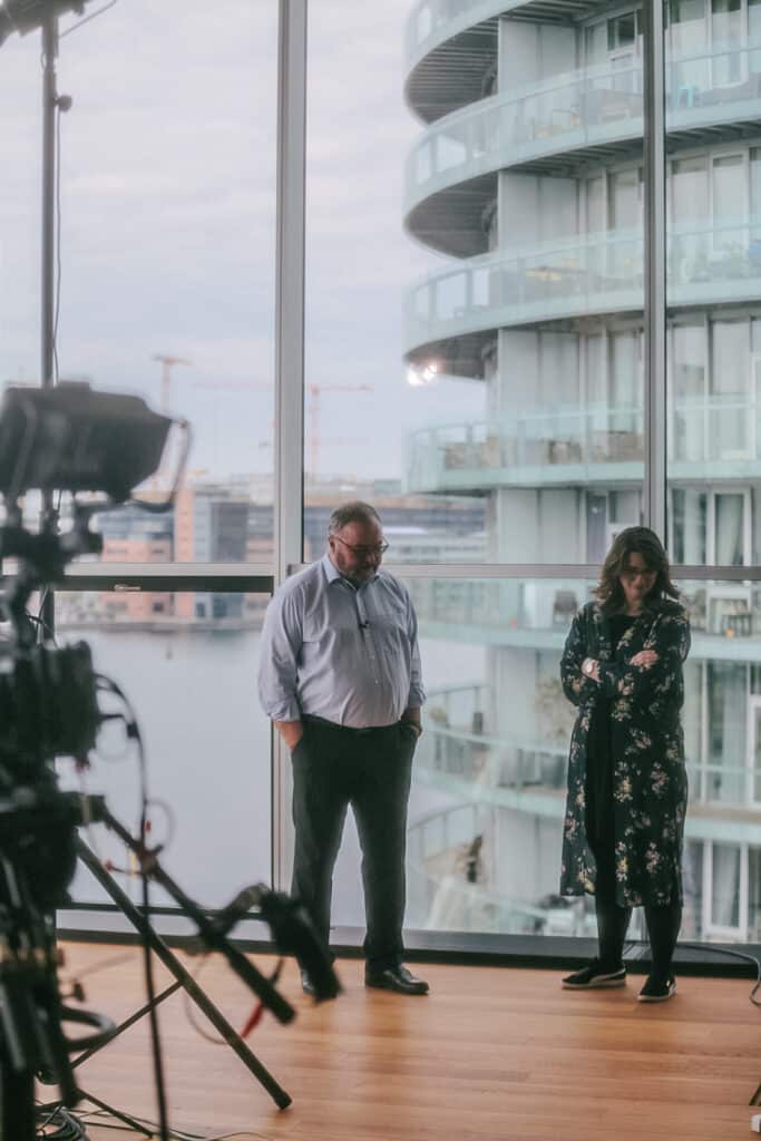En mand og en kvinde venter foran panoramavinduer ud mod Københavns kanaler. I forgrunden står et videokamera på stativ.