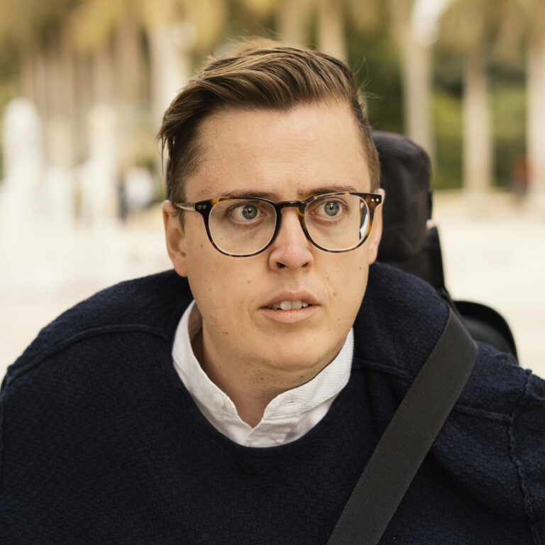 Portrætfoto: Rasmus ser lidt forbi kameraet. Han bærer briller og en mørkeblå trøje over en hvid skjorte. I baggrunden ses slørede træer i varme farver.