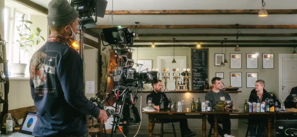 En kameramand filmer en gruppe mennesker i samtale i Mikkeller's bar. Der står et udvalg af flaskeøl foran dem.