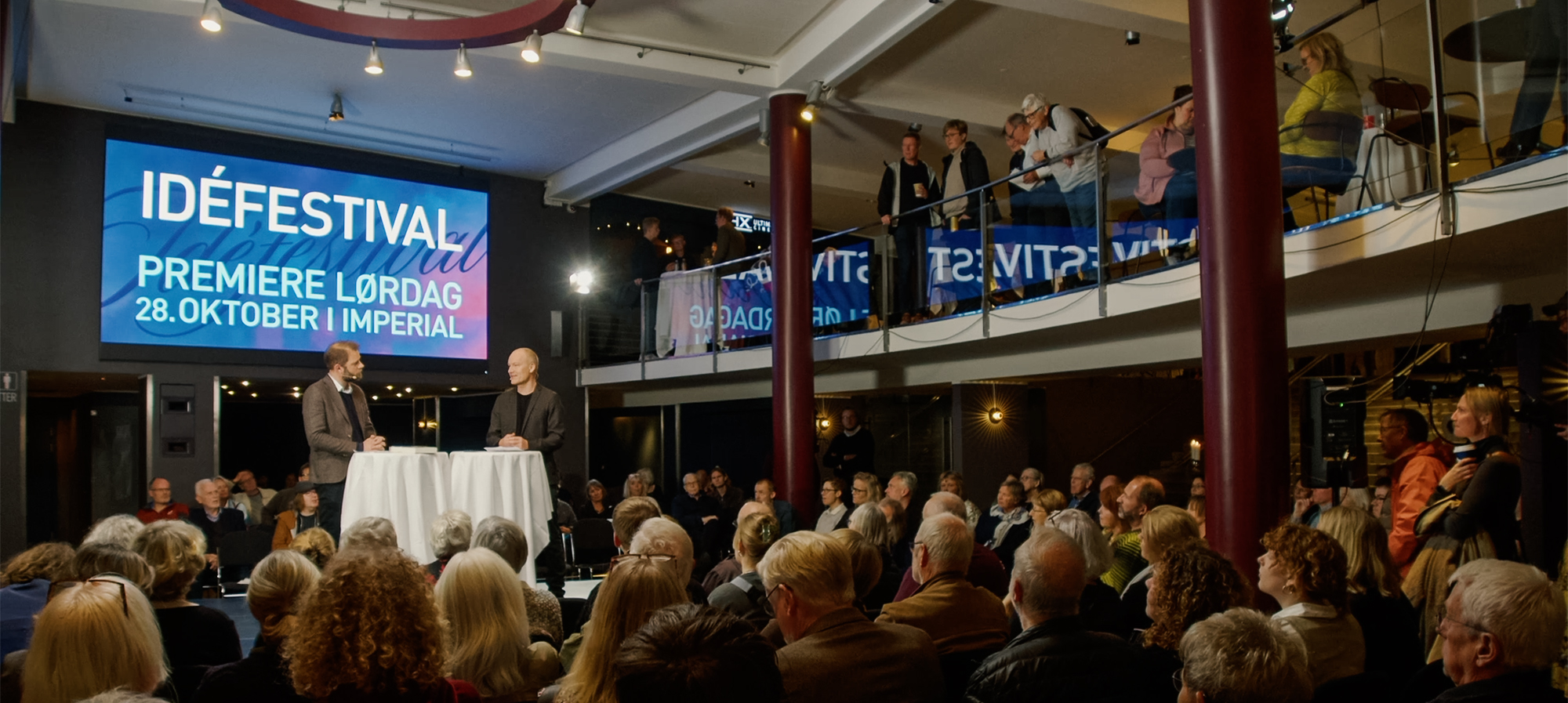 Billedet viser et velbesøgt arrangement til "IDÉFESTIVAL", som fandt sted lørdag den 28. oktober i Imperial. To talere står i centrum for opmærksomheden på et podium i forgrunden, med en engageret forsamling af tilhørere i forskellige aldre, der lytter opmærksomt. En stor skærm i baggrunden fremviser festivalens navn og dato, hvilket bidrager til arrangementets festlige atmosfære. Over publikum er der en balkon, hvor flere står og ser ned på scenen. Stedets arkitektoniske detaljer og belysning skaber en indbydende og kulturelt fyldt atmosfære, som passer til festivalens ånd.